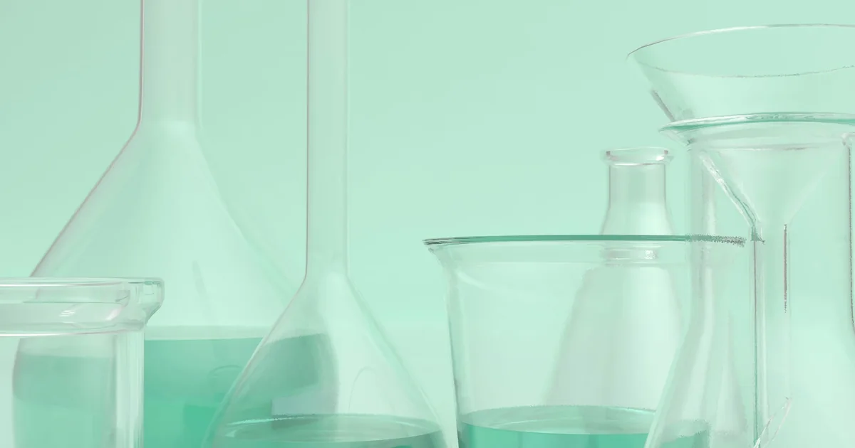 Vidraria de laboratório — balões volumétricos, béqueres e funil — com líquido verde-claro, evocando o estudo dos ativos cosméticos.